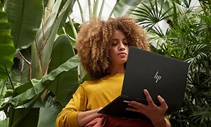 Kvinde står under nogle tropiske planter og holder en HP Envy PC