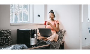 En kvinde med et headset foran sin bærbare computer og ved siden af sin kaffemaskine