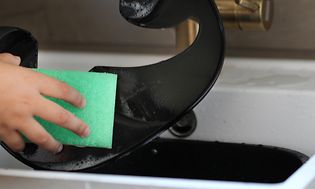 En person rengør airfryerelement med en grøn opvaskesvamp i en køkkenvask